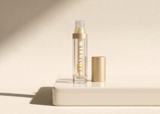 Luxette Velvet Lip Gloss in Clear, gold-capped glass tube standing on a cream pedestal in warm sunlight